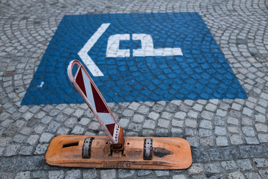 A dedicated parking space for disabled access, marked with a blue square sign featuring a white wheelchair symbol, is painted onto a cobbled pavement surface. An orange temporary traffic barrier with